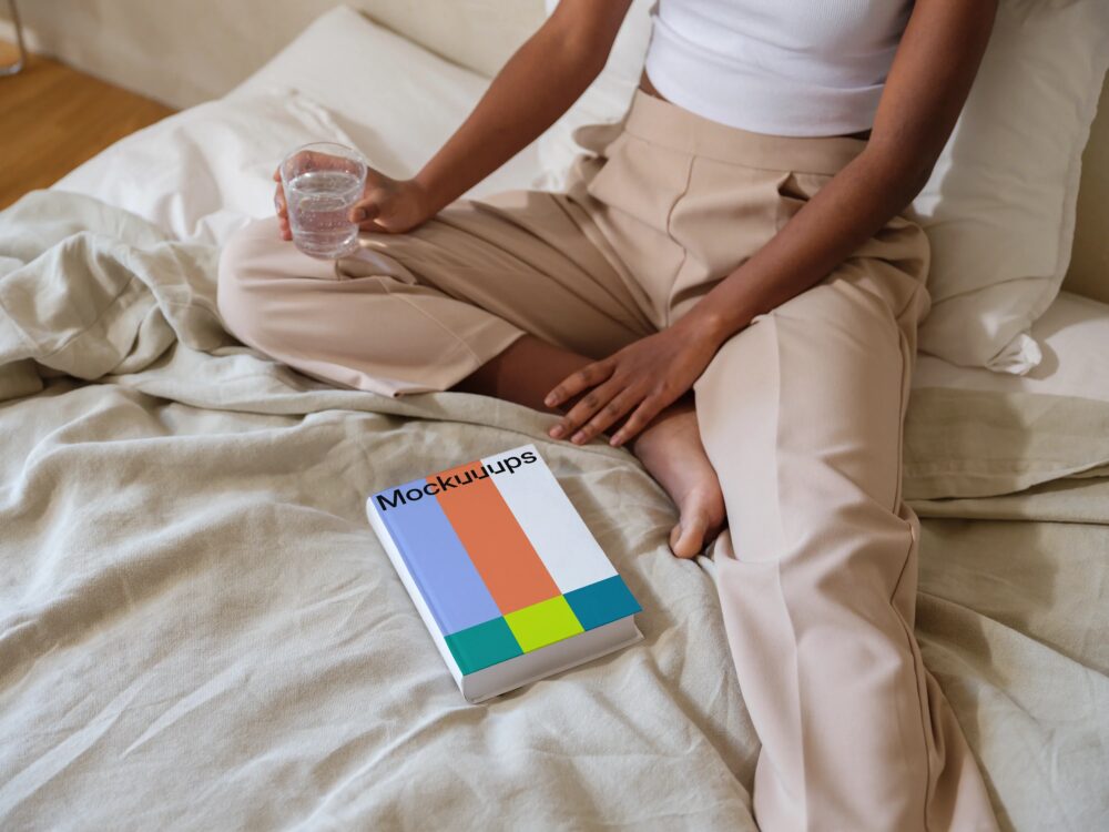 Book mockup on a cozy bed with a person holding a glass of water